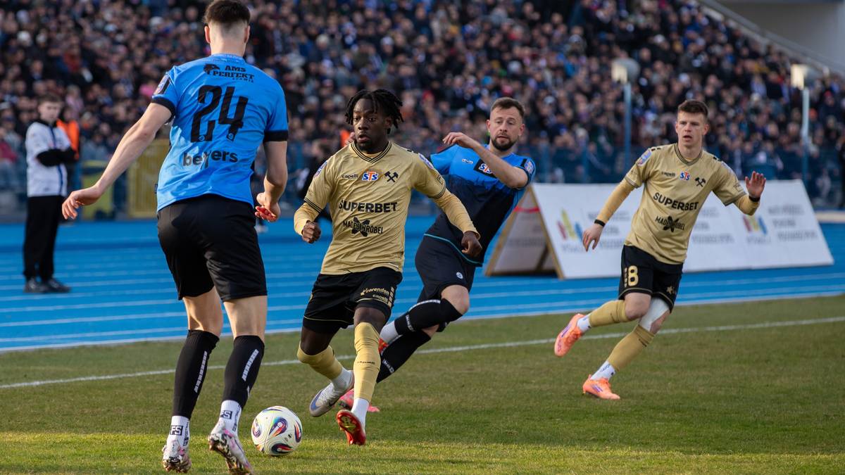 Czterech piłkarzy rywalizuje o piłkę na boisku, w tle trybuny zapełnione kibicami.