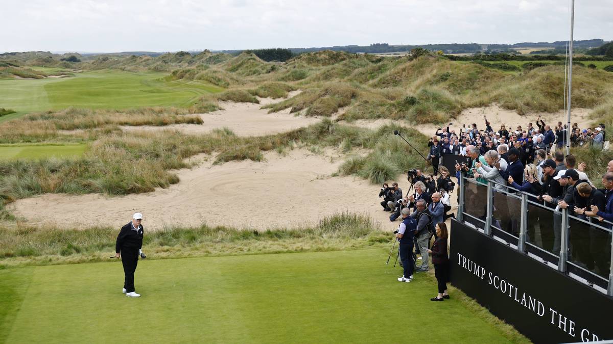 Gracz w białej czapce stoi na zielonym polu golfowym, obok niego widać piaszczyste wydmy porośnięte trawą i grupę ludzi stojących za barierą.