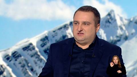 Grzegorz Michalewski, mężczyzna w ciemnym garniturze, m&oacute;wi przed kamerą na tle ośnieżonych g&oacute;r. W prawym dolnym rogu znajduje się małe okno z tłumaczką języka migowego.
