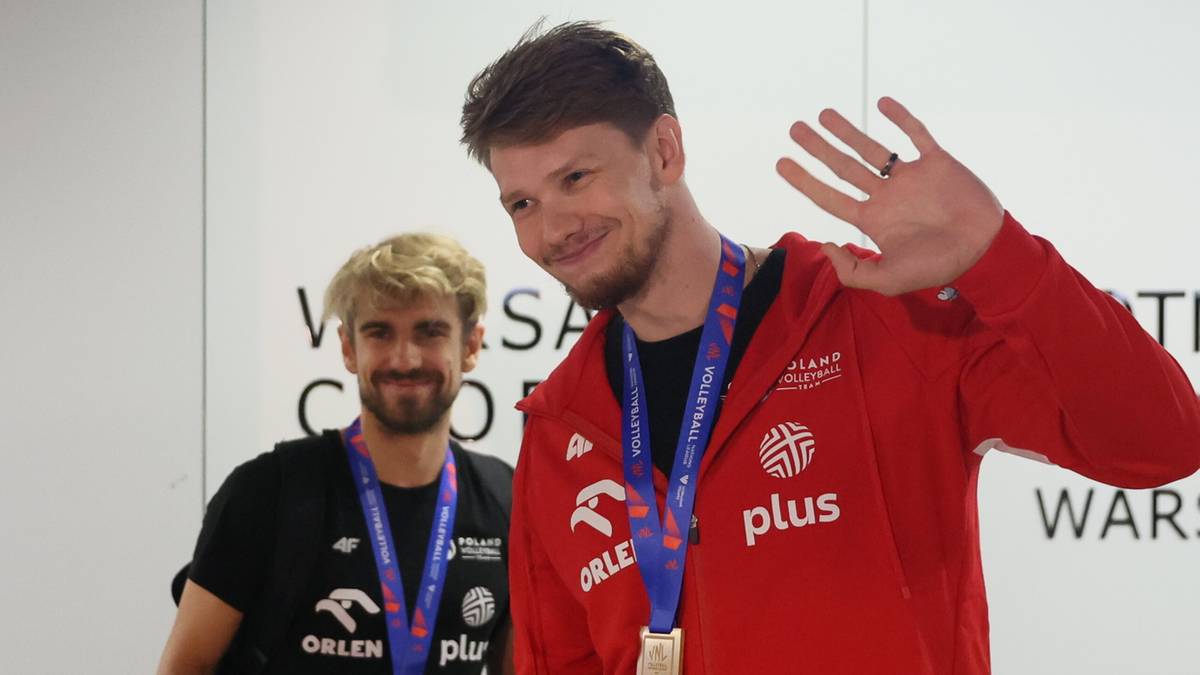 Dwóch polskich siatkarzy ubranych w stroje sportowe, z medalami na szyjach, jeden z nich uśmiecha się i macha do obiektywu.