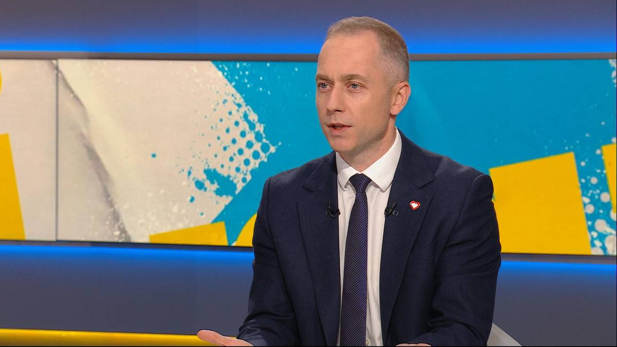 Mężczyzna w eleganckim granatowym garniturze wypowiada się w studio telewizyjnym na tle niebiesko-ż&oacute;łtej scenografii.
