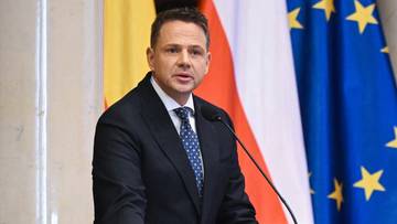 Mężczyzna w garniturze przemawia przy m&oacute;wnicy na tle flagi Polski i Unii Europejskiej.