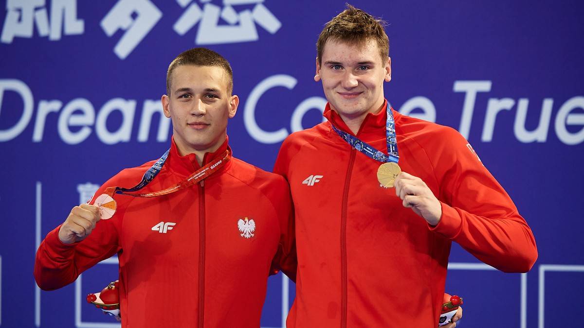 Dwóch młodych mężczyzn w czerwonych strojach sportowych, obaj z medalami na szyi, uśmiechają się do zdjęcia.