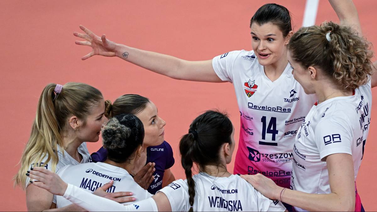 Grupa siatkarek w białych strojach dyskutuje na boisku, jedna z nich gestykuluje ręką.
