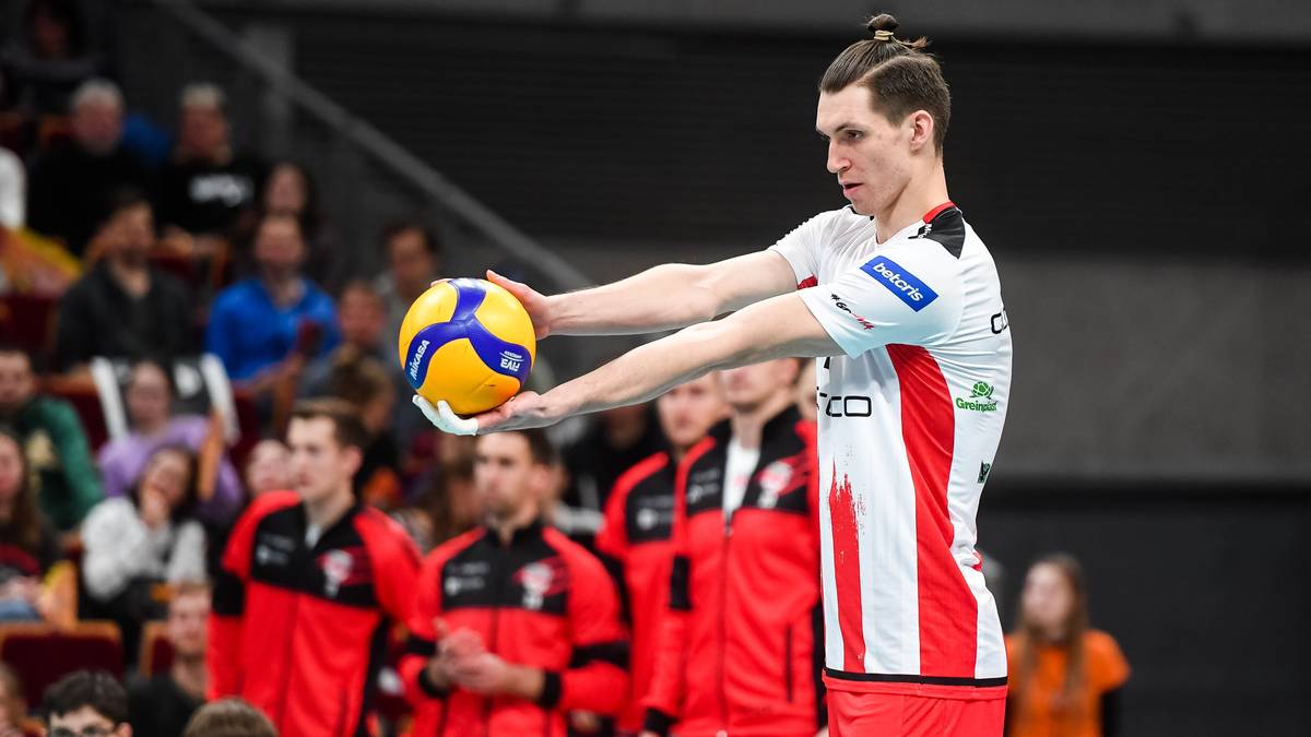 Siatkarz Maciej Muzaj trzymający piłkę do siatk&oacute;wki, ubrany w biało-czerwoną koszulkę sportową, na tle publiczności.
