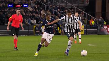 Piłkarze walczący o piłkę na boisku podczas meczu piłki nożnej. Jeden z nich nosi granatowo-białą koszulkę, drugi biało-czarne pasy. W tle widać trybuny z kibicami i sędziego.