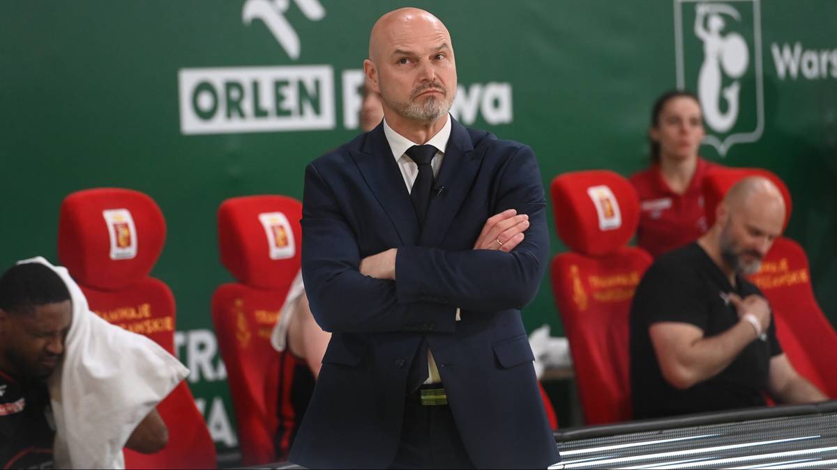 Trener koszykówki w garniturze stoi z założonymi rękami, patrząc w stronę obiektywu. W tle widoczni są inni członkowie drużyny i kolorowe siedzenia.