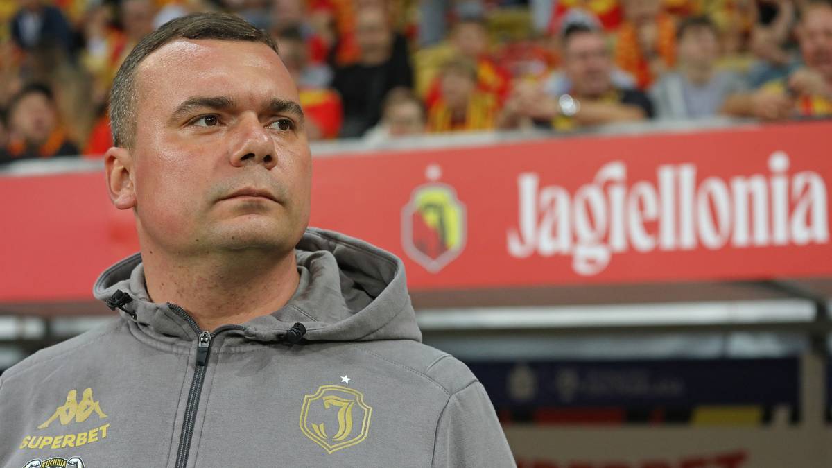 Mężczyzna w szarej bluzie z kapturem z logo klubu sportowego, stojący na tle stadionu z banerem "Jagiellonia". Ma poważny wyraz twarzy.