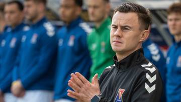 Trener drużyny piłkarskiej z dłońmi złożonymi w geście modlitwy lub koncentracji, w tle widoczni zawodnicy w niebieskich strojach.