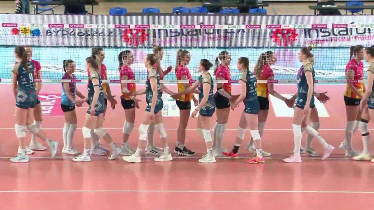 Dwie drużyny siatkarek, jedna w niebiesko-granatowych strojach, druga w różowych, stoją naprzeciwko siebie na parkiecie hali sportowej.