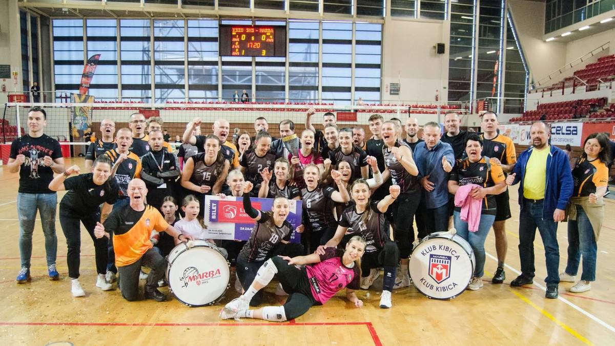 Drużyna siatkarska w komplecie z zawodniczkami, sztabem szkoleniowym i osobami towarzyszącymi, pozuje do wspólnego zdjęcia w hali sportowej, trzymając trofea i okrzyki radości.
