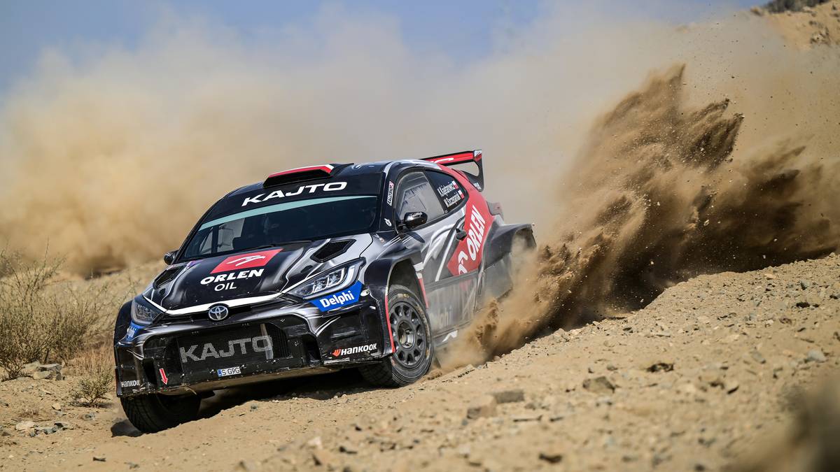 Samochód rajdowy Toyota GR Yaris z numerem 18, z logotypami "KAJTO" i "ORLEN", pokonuje pustynny odcinek specjalny, wzbijając chmurę piasku.