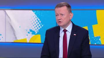 Mariusz Błaszczak w garniturze i krawacie, udziela wywiadu na tle ekranu z abstrakcyjnym wzorem.