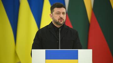 Wołodymyr Zełenski przemawia za m&oacute;wnicą z flagą Ukrainy.