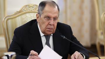 Mężczyzna w garniturze siedzi przy stole z dokumentami i mikrofonem.