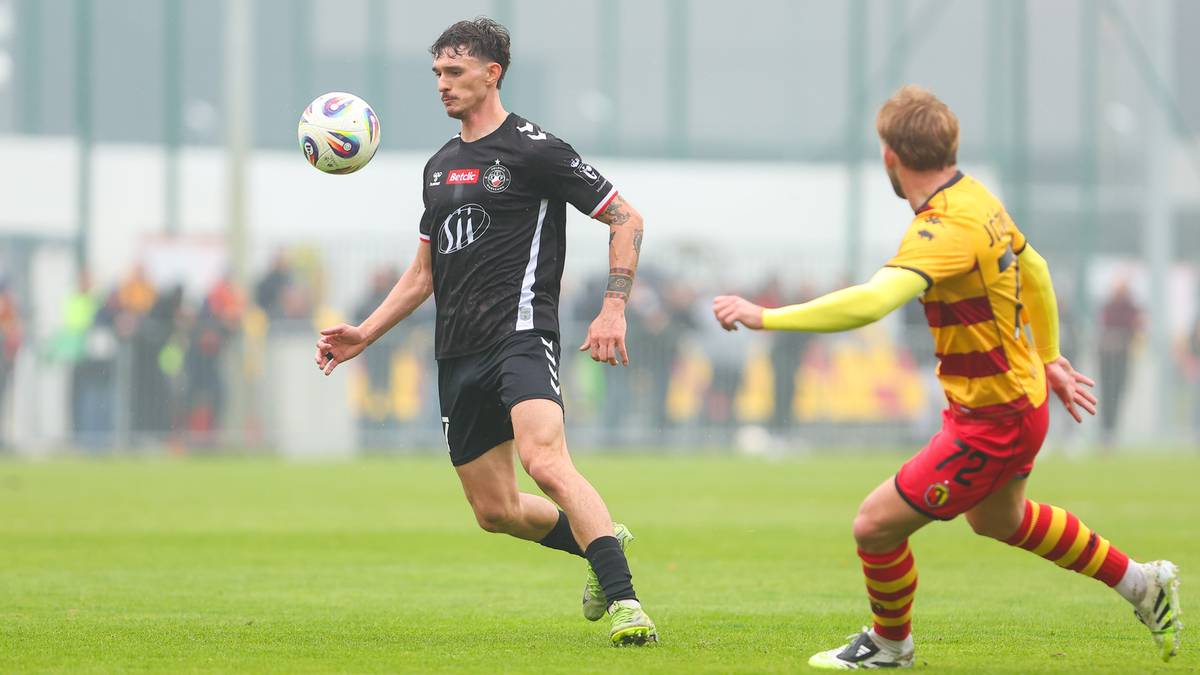 Dw&oacute;ch piłkarzy na zielonej murawie, jeden w czarno-białej koszulce kontroluje piłkę, drugi w ż&oacute;łto-czerwonej koszulce biegnie za nim.