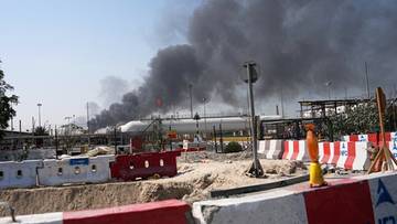 Kłęby czarnego dymu unoszące się nad budynkami, widoczne zza tymczasowych barierek budowlanych.