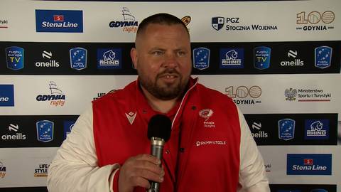 Mężczyzna w białej kurtce z czerwonymi elementami, trzymający mikrofon i m&oacute;wiący.