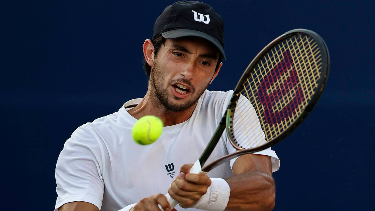 Mężczyzna w czarnej czapce z daszkiem i białej koszulce uderza piłkę tenisową rakietą.