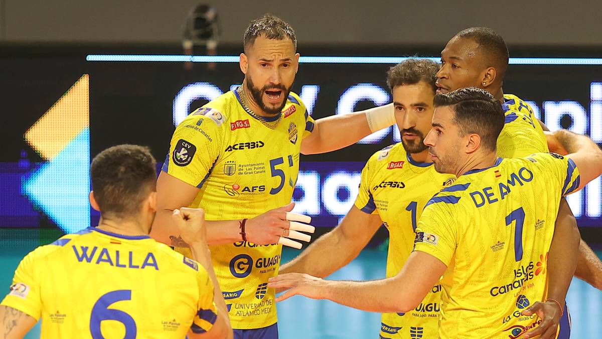 Pięciu mężczyzn w ż&oacute;łtych koszulkach i niebieskich spodenkach świętuje na boisku do siatk&oacute;wki.