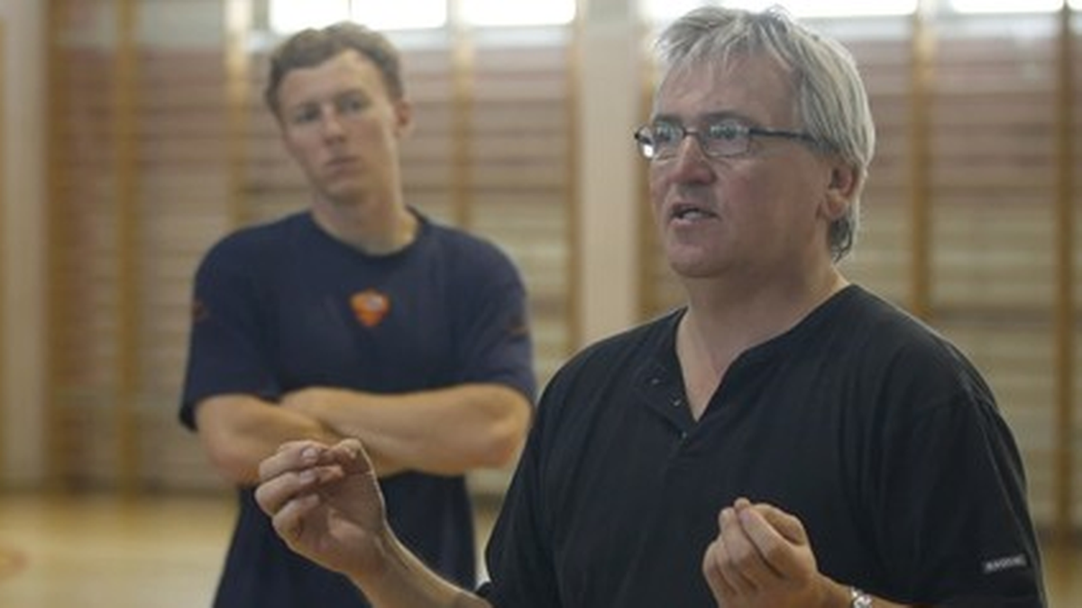 Mężczyzna w średnim wieku, trener, mówi energicznie i gestykuluje. W tle stoi młodszy mężczyzna, słuchając.