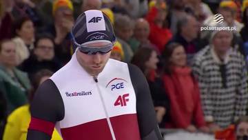 Łyżwiarz szybki w czapce z daszkiem i okularach ochronnych, ubrany w biało-czerwony str&oacute;j sportowy z logotypami PGE i 4F, z widownią na trybunach w tle.