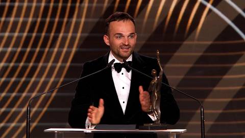 Bartosz Zmarzlik w czarnym smokingu z białą koszulą i muszką, trzymający statuetkę przy mównicy, z rękami lekko uniesionymi.
