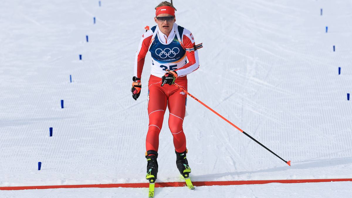 Biathlonista w czerwonym stroju z karabinem na plecach biegnie po śniegu na nartach, z numerem 35 i logo igrzysk olimpijskich na piersi.