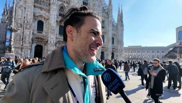 Mężczyzna z włosami związanymi w kok, w brązowym płaszczu i niebieskiej kurtce, udziela wywiadu przed wielką kamienną budowlą.