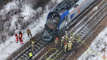 Widok z g&oacute;ry na miejsce wypadku kolejowego, gdzie pociąg zderzył się z samochodem osobowym, kt&oacute;ry zapalił się. Na torach widoczni są strażacy.