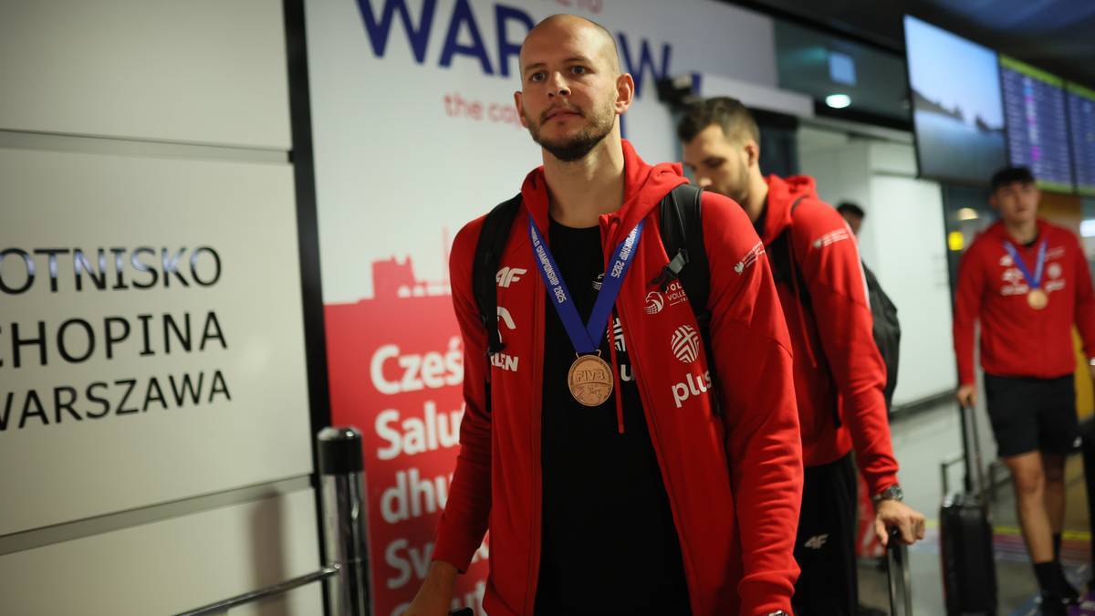 Skoczek narciarski w czerwonej bluzie z kapturem i medalem na szyi idzie przez lotnisko, trzymając plecak na ramieniu.