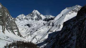 Ośnieżony szczyt g&oacute;rski z lodowcem i skalistymi zboczami w tle, z lasem iglastym na pierwszym planie pod niebieskim niebem.