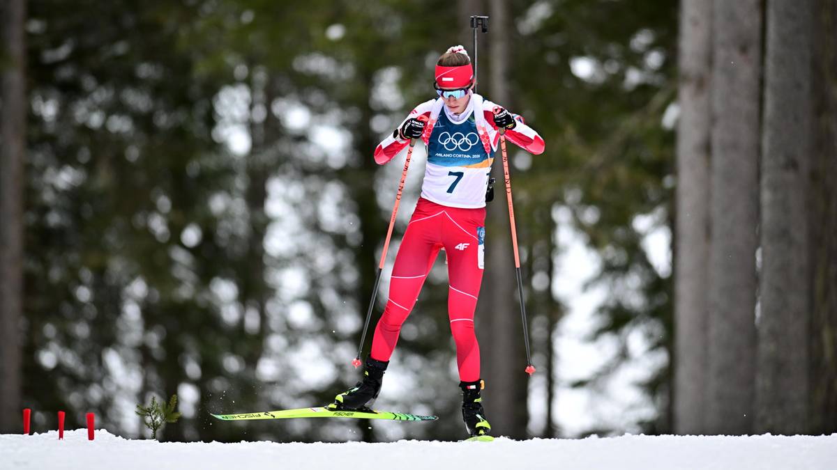 Biathlonistka w czerwonym stroju z numerem 7 na piersi, na nartach z kijami narciarskimi, na tle drzew i śniegu.