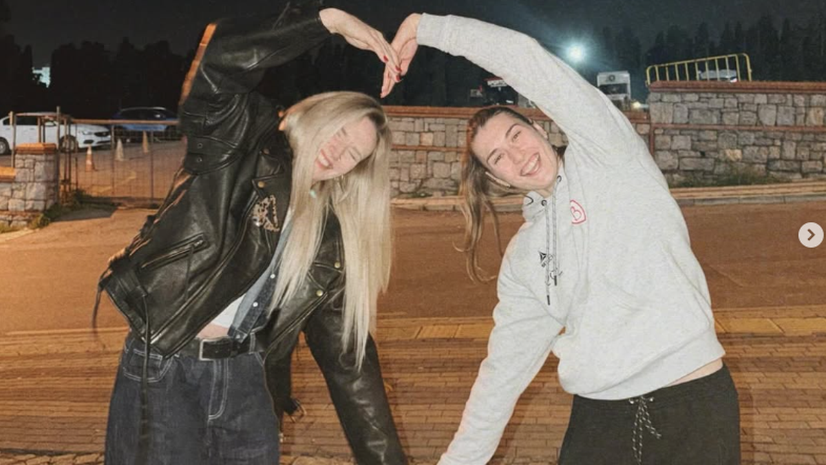 Dwie młode kobiety stojące na zewnątrz, tworzące serce z dłoni nad głowami.