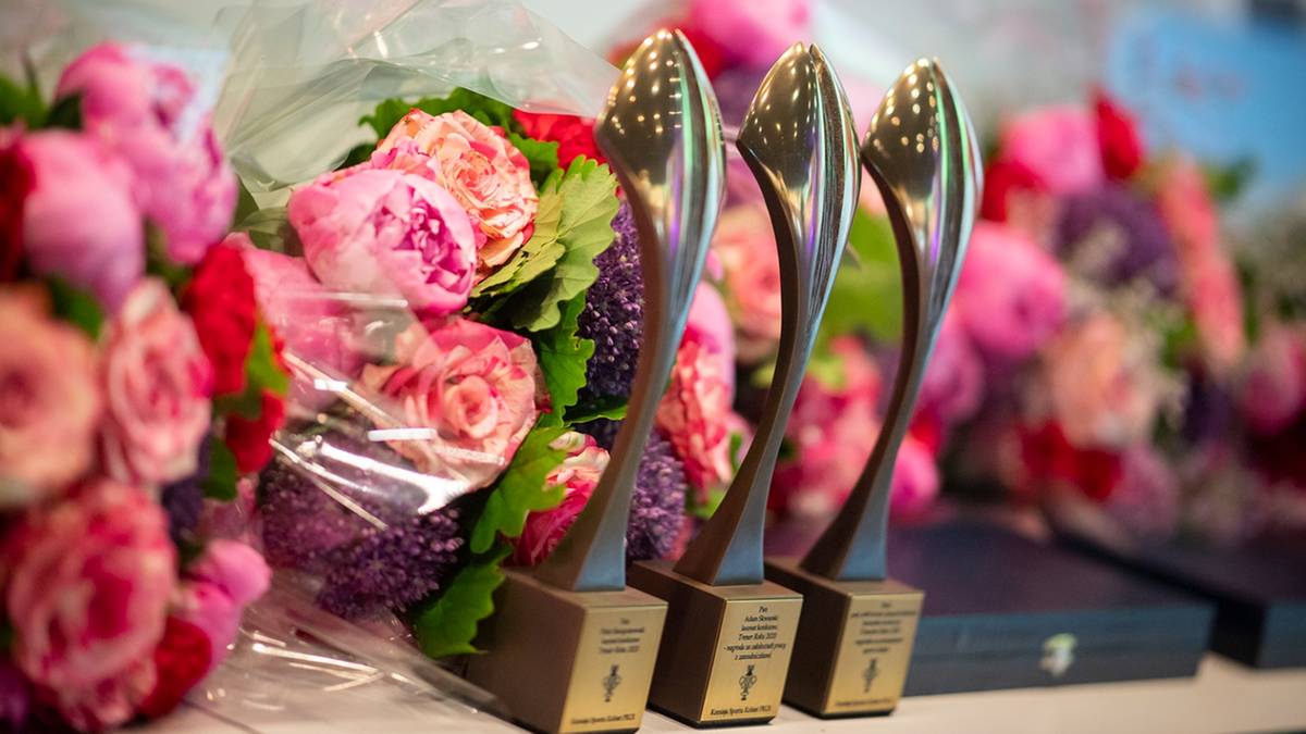 Trzy srebrne statuetki konkursowe stoją obok bukiet&oacute;w r&oacute;żowych i fioletowych kwiat&oacute;w. Tło jest nieostre i przedstawia więcej kwiat&oacute;w oraz ciemny, prostokątny przedmiot.
