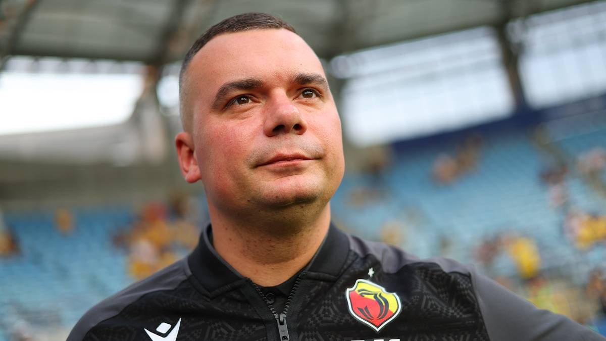 Portret mężczyzny w stroju sportowym z herbem klubu na piersi, na tle trybun stadionu.
