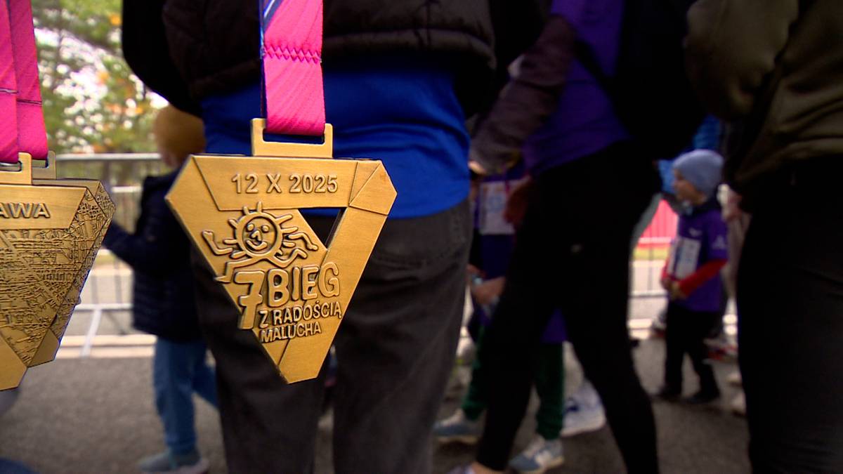 Złoty medal z różową wstążką, na którym widnieje napis "12 X 2025 7. BIEG Z RADOŚCIĄ". Medal przypomina kształtem trójkąt z wyciętym kołem w środku, w którym znajduje się symbol słońca i dziecka. W tle widoczni są uczestnicy biegu.